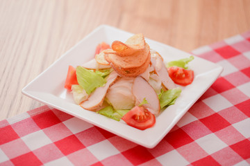 Plate of traditional Caesar Salad with chicken and salad on light wooden table with red plaid tablecloth.