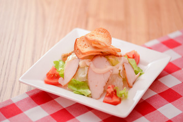 Plate of traditional Caesar Salad with chicken and salad on light wooden table with red plaid tablecloth.