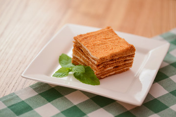 A slice of layered vanilla honey cake on a white plate with a mint leaf on wooden table with a plaid table cloth