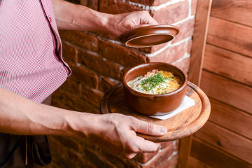 Obraz na płótnie Canvas Waiter holds a bowl with potato gratin with cream and cheese and mushrooms in a restaurant. Waiter in a plaid uniform.