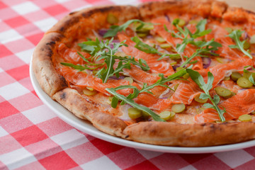 Delicious Italian pizza with salmon fish served on white plate on rustic wooden table with plaid tablecloth.