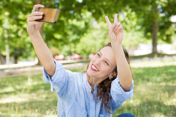 Smiling young woman showing peace sign while taking selfie. Cheerful lady holding phone in outstretched arm and gesturing. Technology, self portrait concept