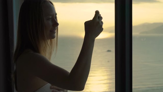 A young woman in an apartment with a panoramic view on the coast and sunrise. She makes a photo of a beautiful sunrise