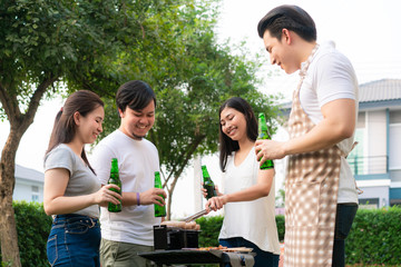 Asian man cooking barbeque grill and sausage for a group of friends to eat party in garden at home. group of friends having outdoor garden barbecue laughing with alcoholic beer drinks.