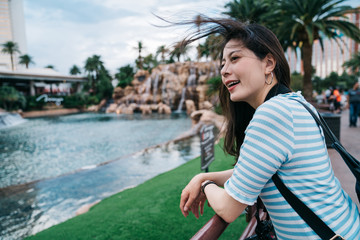 Fototapeta na wymiar side view of happy young girl backpacker stands at fountain in famous hotel in las vegas. smiling asian woman traveler leaning on railing sightseeing beautiful water dance show in nature summer park