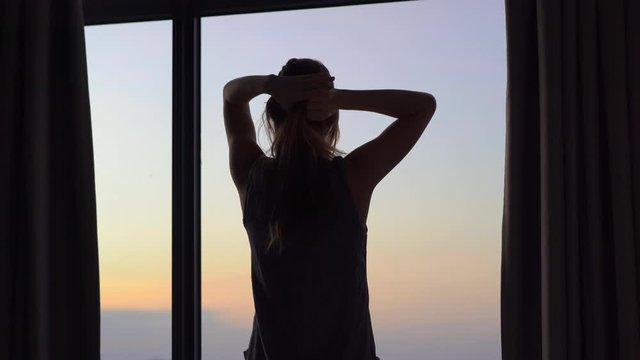 A young woman in an apartment with a panoramic view on the coast and sunrise. She opens window curtains