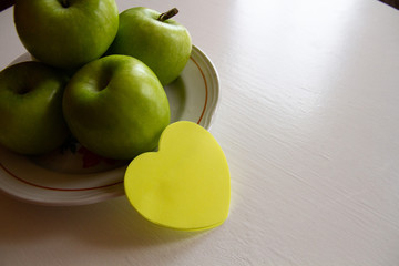 On the white table is a plate with green apples, next to a yellow sticker for the inscription