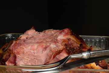 Close-up of an appetizing smoked pork leg ham leftovers on a square glassware and partial view of a silver cutting fork and knife, with black background 