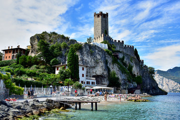 Malcesine, Italia - Scaliger Castle, a medieval lakeside castle with fortified walls, on the shores of Lake Garda, located on a cliff, green bushes and trees below, a beach.