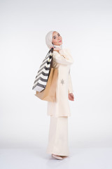 A beautiful Muslim female model in a Asian traditional dress kurung Pahang carrying shopping bags isolated on white background. Eidul fitri festive preparation shopping concept. Full length portrait.