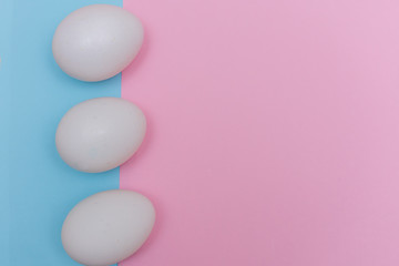 Three Easter holiday flat lay with white egg on a solid light blue, and pink pastel background with copy space.