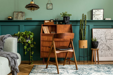 Stylish interior design with retro wooden cabinet, chair, gray sofa, plants, pendant lamp, decoratnion, maps, stool and elegant personal accessories. Modern retro concept of home office space.