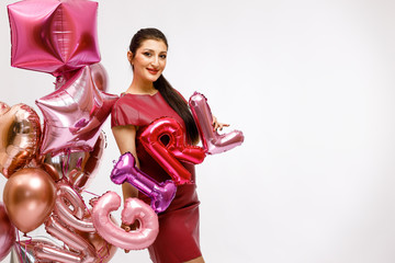 A young beautiful brunette girl in a pink leather dress is holding a balloon with the inscription "GIRL" for Valentine's day, hen party or baby shower on a white background with bright helium balloons