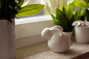 Ceramic easter bunny on windowsill with green leaves decoration and blooming daisies in flowerpot. Waiting for spring, background with copy space, easter home decoration.