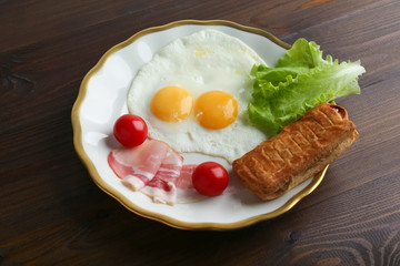 fried eggs served with bacon, cherry tomatoes, lettuce and a puff pastry on a plate on dark wooden background