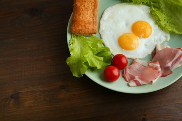 fried eggs with bacon, cherry tomatoes, lettuce and a puff pastry on a plate on dark wooden background