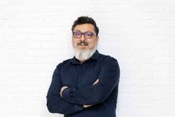 Relaxed multiethnic senior business man wearing glasses with trendy beard and arms folded against white brick wall - 322956795