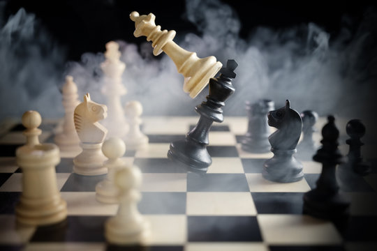 chess queen beats king between other pieces on the chessboard, much smoke over the battle,  against a dark background, concept for aggression, success and competition between teams