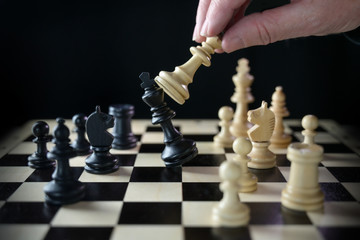 Human hand is playing chess and defeats the king, checkmate on the chessboard against a black...