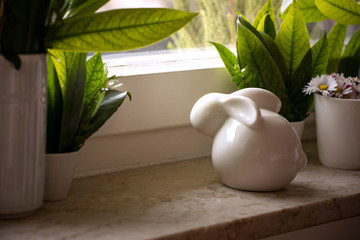 Ceramic easter bunny on windowsill with green leaves decoration and blooming daisies in flowerpot. Waiting for spring, background with copy space, easter home decoration.