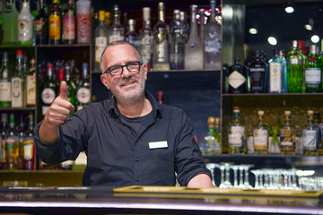 Portrait of elderly professional bartender in eyeglases show hand OK in workplace closeup. High level and quality service. Evening time. The concept of service.