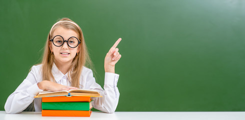 Young girl wearing funny eyeglasses sits with books and points away on empty green chalkboard. Empty space for text