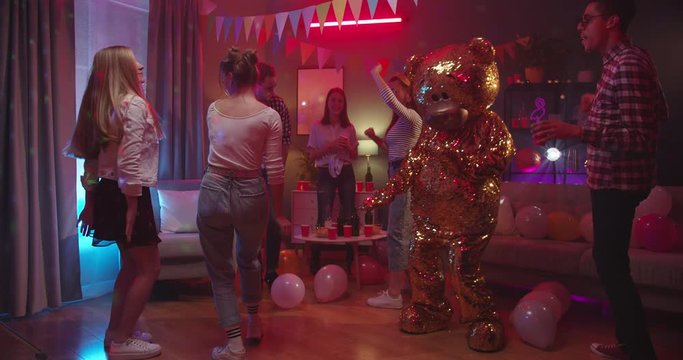 Big funny golden grown doll of teddy bear dancing at the home party among young happy guys and girls in the decorated room.