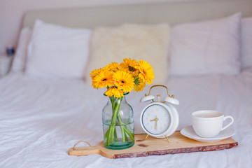 A bouquet of gerberas in a glass jar, a cup of cappuccino, a white alarm clock on a wooden stand stand on the bed. Breakfast in bed.