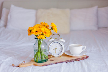 A bouquet of gerberas in a glass jar, a cup of cappuccino, a white alarm clock on a wooden stand stand on the bed. Breakfast in bed.
