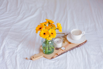 A bouquet of gerberas in a glass jar, a cup of cappuccino, a clock, a candle on a wooden stand are on the bed. Breakfast in bed.