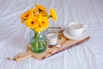 A bouquet of gerberas in a glass jar, a cup of cappuccino, a clock, a candle on a wooden stand are on the bed. Breakfast in bed.