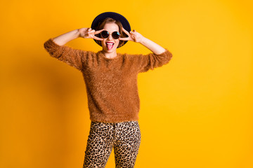 Photo of beautiful funny lady hold fingers v-sign symbol near eyes good mood stick tongue out mouth wear sweater leopard pants sun specs retro cap isolated yellow color background