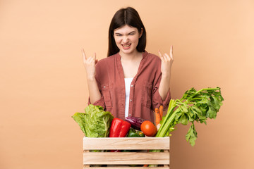 Young farmer girl with freshly picked vegetables in a box making rock gesture