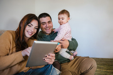 Portrait of a family on the sofa at home while looking at the tablet - Father, mother and one year old daughter have fun together - Intimacy moment - Copy space