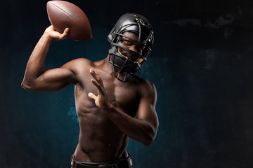 portrait photo of dark-skinned young man with nude torso on a dark background he has a rugby helmet on head and holds a rugby ball up in his arm