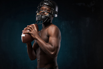 portrait photo of dark-skinned young man with nude torso on a dark background he has a rugby helmet on head, holds a rugby ball in his arms