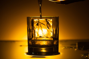 vodka pouring into transparent glass with ice cube in dark with warm back light