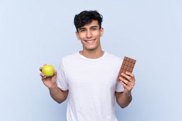 Young Argentinian man over isolated blue background taking a chocolate tablet in one hand and an...