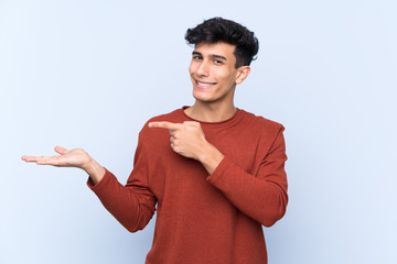 Young Argentinian man over isolated blue background holding copyspace imaginary on the palm to...