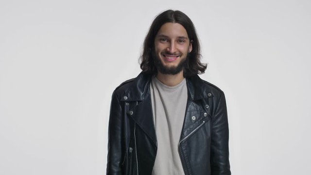 A smiling young man with long hair in a black leather jacket is sending an air kiss to the camera isolated over white background