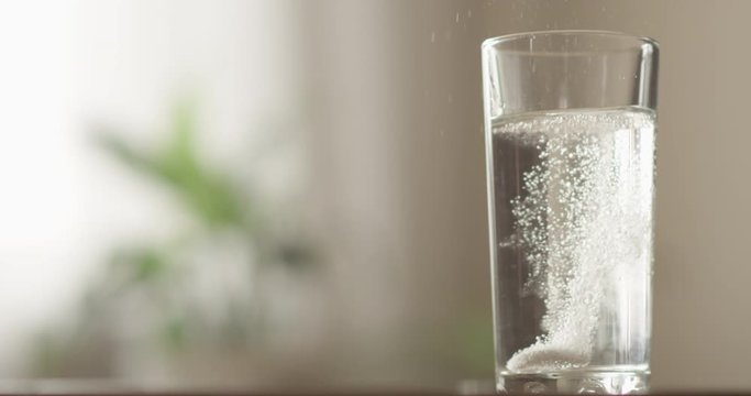 Soluble Effervescent Aspirin Tablet Dropped in a Glass and Dissolving in Fizzy Water and Hand Picking Up Headache Medicine Indoors with a Green Plant on the Background a Concept of Health Care Shot on
