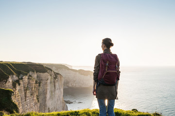 Young woman traveler with backpack looking at sea in Normandy, France over beautiful cliffs background. Travel, active lifestyle and summer holiday concept