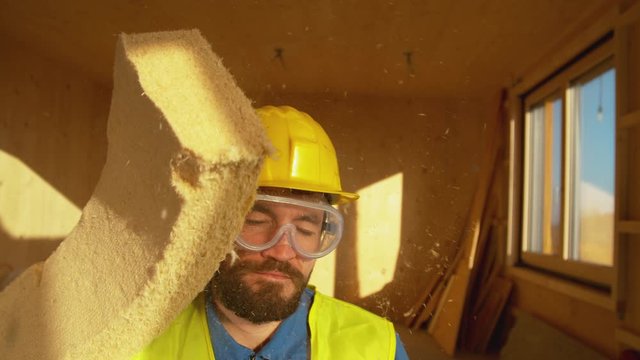 SLOW MOTION, CLOSE UP, PORTRAIT: Bearded construction worker wearing a yellow hard hat gets struck by a piece of foam. Funny shot of a dusty piece of insulation falling on the unsuspecting builder.