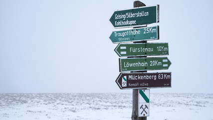 Several hiking signs leading in 2 different directions in fron of a coudy and foggy winter scene  in the background.