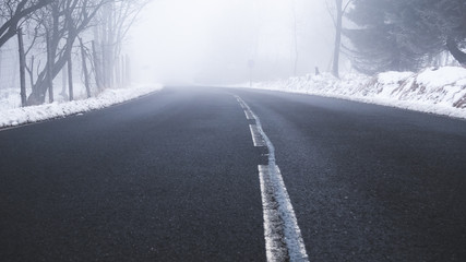 An asphalt road in a rural setting leading into thick fog in the distance on a snowy winters day.