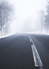 An asphalt road in a rural setting leading into thick fog in the distance on a snowy winters day.