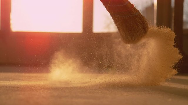 SLOW MOTION, LENS FLARE, CLOSE UP, LOW ANGLE: Dust gets swept up into air as an unrecognizable person cleans a construction site. Contractor sweeps the ground of a construction site with a straw broom
