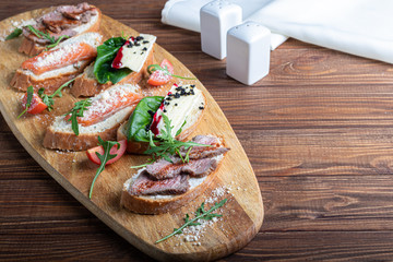 Sandwiches with red fish, beef in suvit and soft cheese. Garnished with slices of microgreen, arugula and tomatoes. On a wooden oval tray.