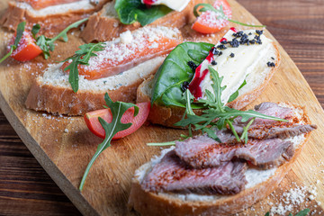 Sandwiches with red fish, beef in suvit and soft cheese. Garnished with slices of microgreen, arugula and tomatoes. On a wooden oval tray.