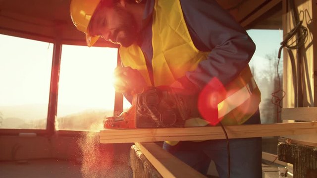 SLOW MOTION, LENS FLARE, CLOSE UP: Golden evening sunbeams shine through the window as the young worker sands a long board. Carpenter working at a construction site buffs a plank on a sunny morning.
